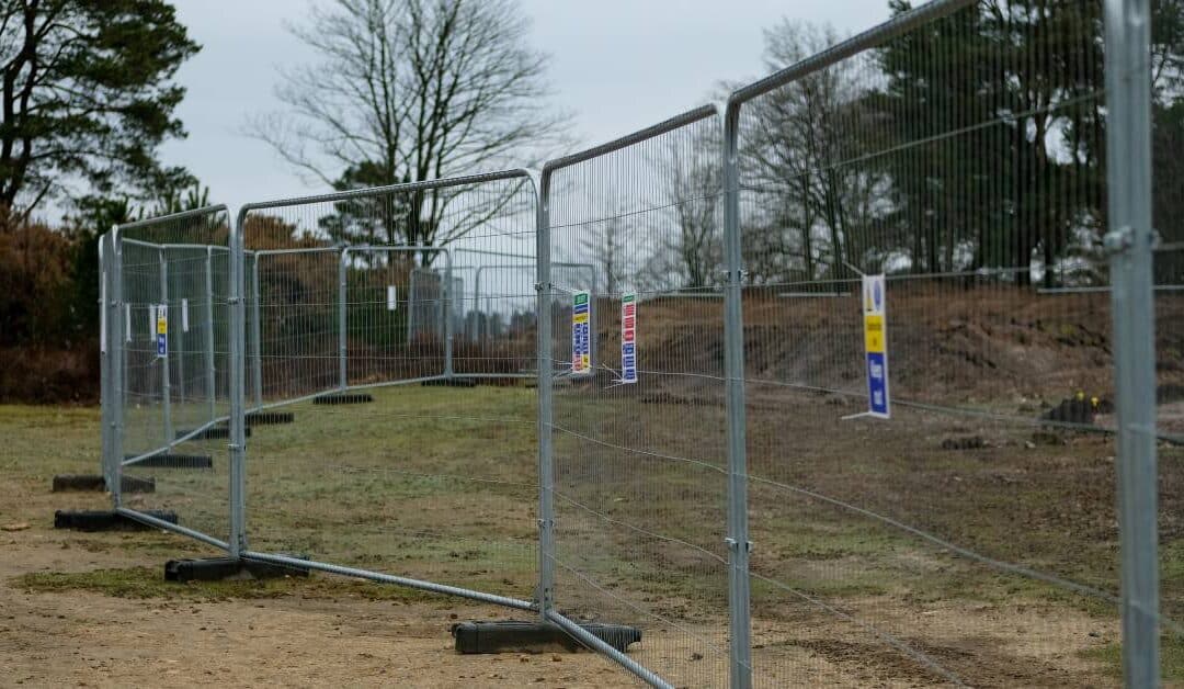 A long line of temporary fence panels cutting through a park. There are a few dirt mounds behind the fence.