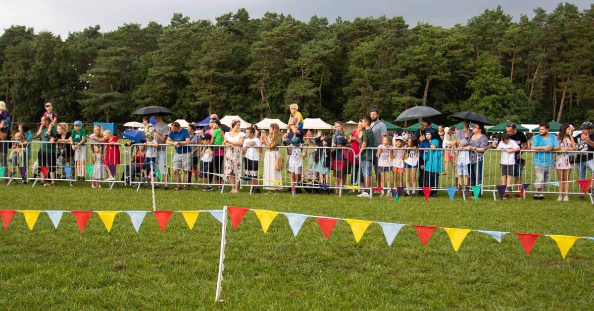 A line of people at an outdoor coordinated event. They are separated by the main event by a crowd control fence.